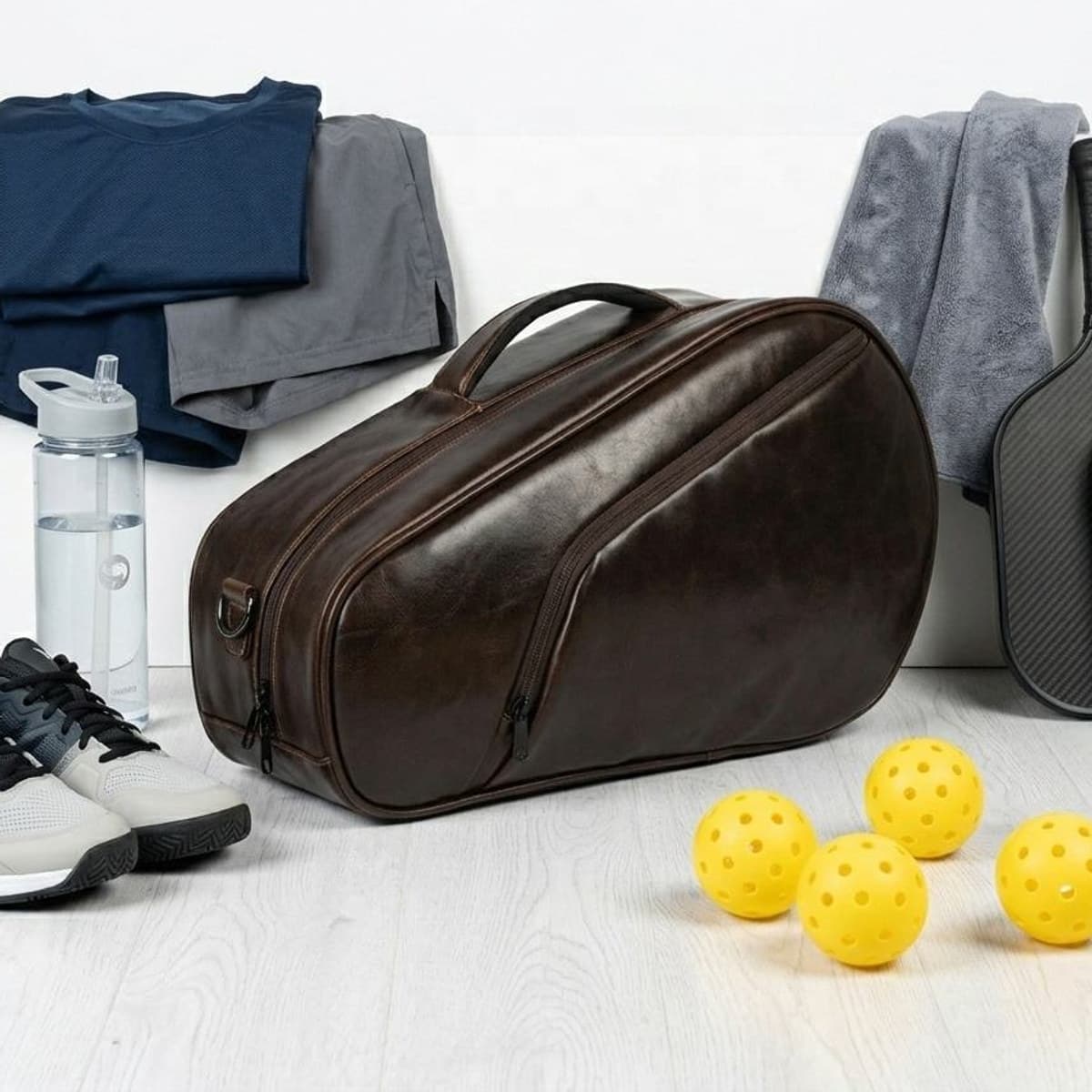 Dark brown PU leather Picklr court tote on a wooden floor beside shoes, a bottle, a towel and pickleballs